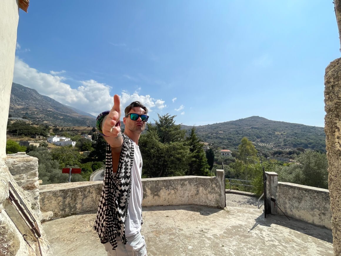 Aiden pointing to the camera in a Greek buildings with mountains behind