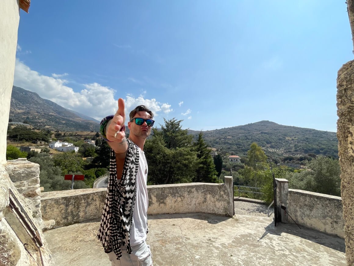 Aiden pointing his fingers to camera at old building in Crete with mountains in background