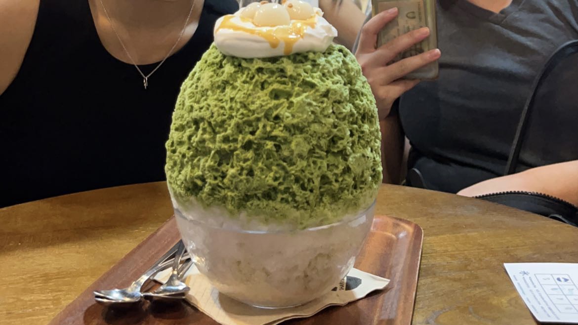 a huge bowl of green shaved ice with a white creamy topping and syrup