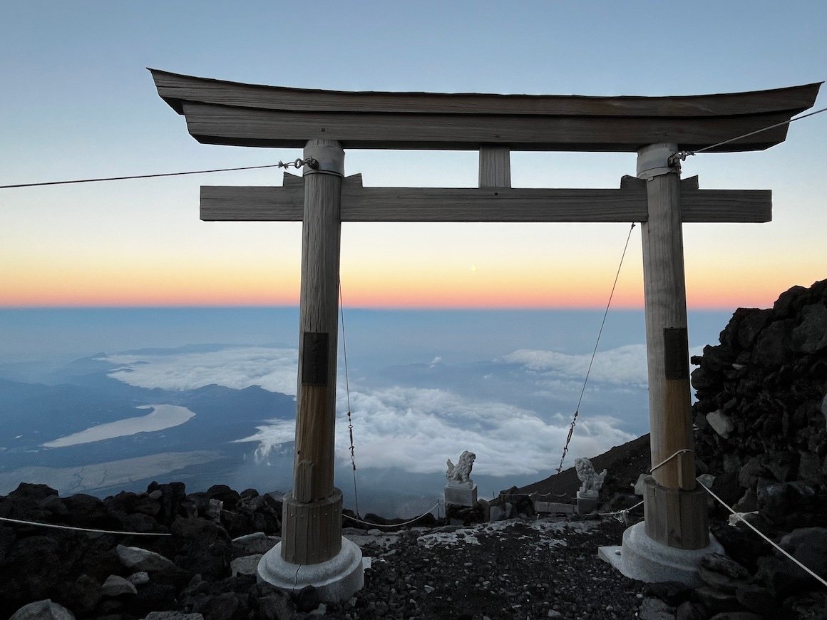 A sunset view of torii gate on a rocky mountaintop in Japan, with Mount Fuji in the background.