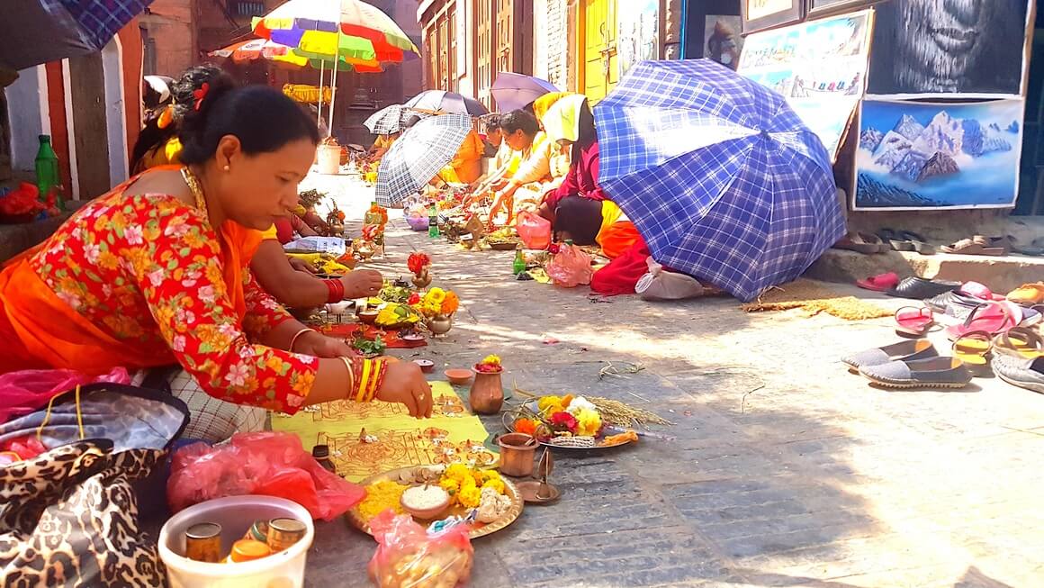 Nepali women in bright clothes sat on the floor outside Hindu temple in bright clothes making plated of offerings of flowers and food.