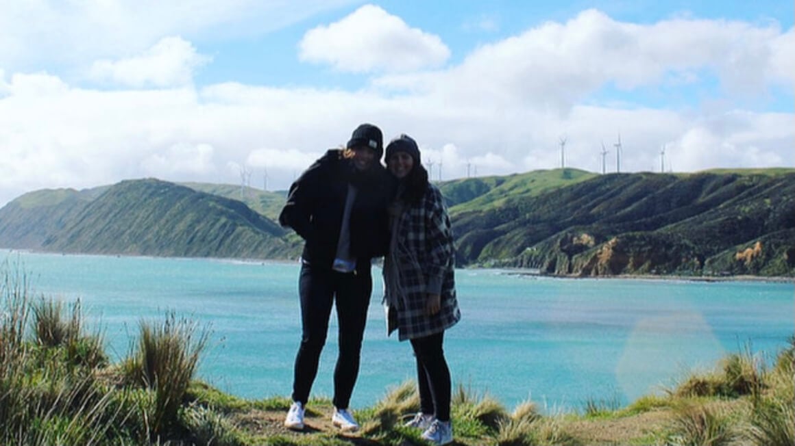 Two girls at Makara Beach with ocean, mountains and windmills behind them.