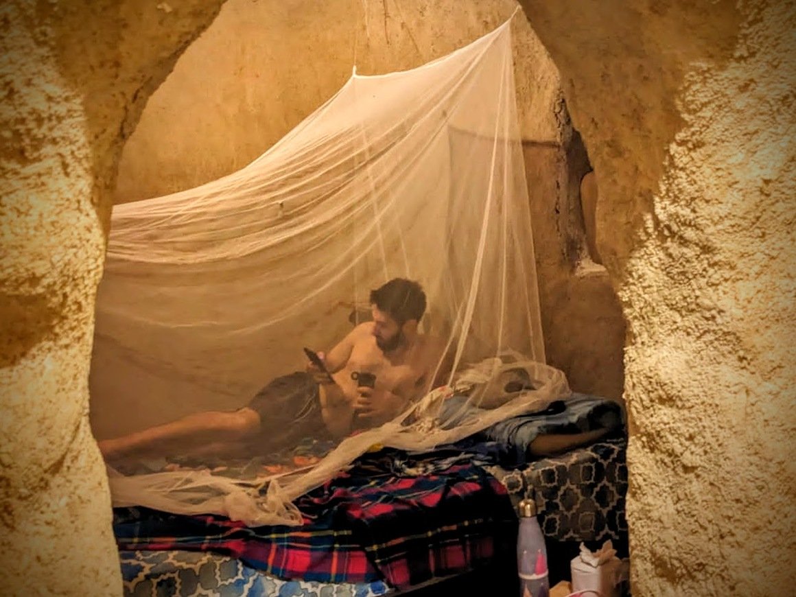 Man laying on a bed looking at a phone inside a mosquito net in a natural building hut