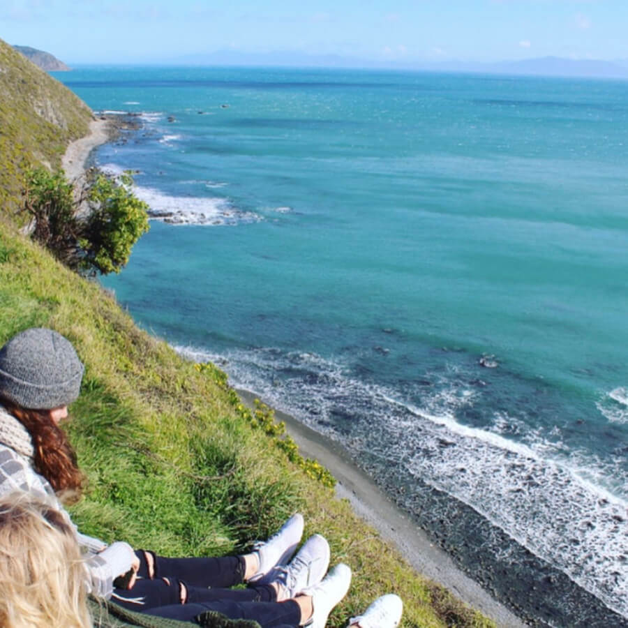 Two girls sitting on top of a hill looking over beauty blue sea.