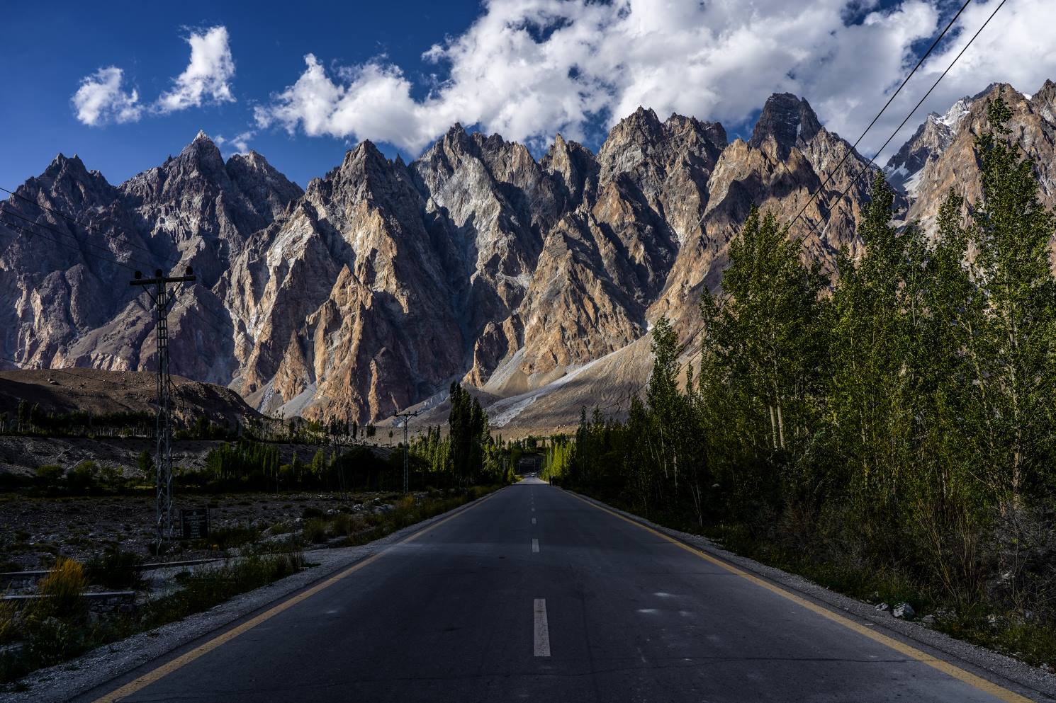 a massive wall of mountains towering over a paved black highway in hunza valley northern pakistan