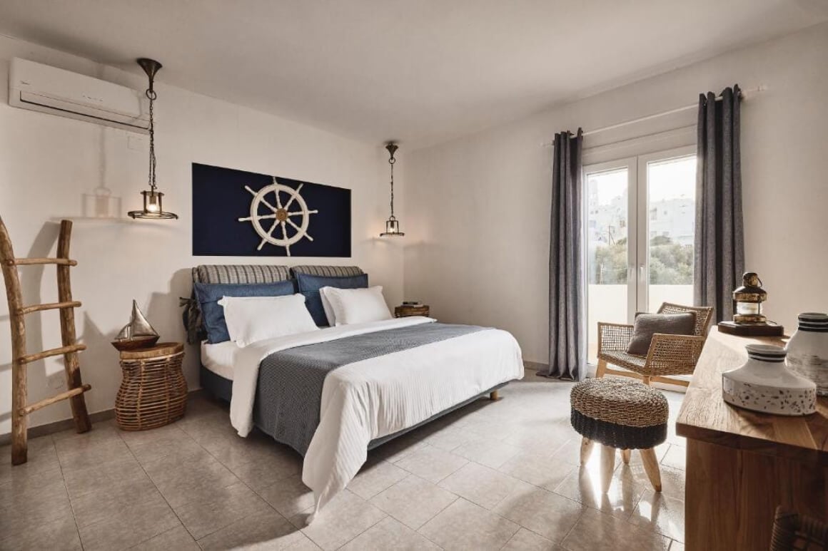 Sailor style bedroom with a queen bed with blue and white sheets. There is a canvas with a picture of a sailing boat steering wheel behind the bed.