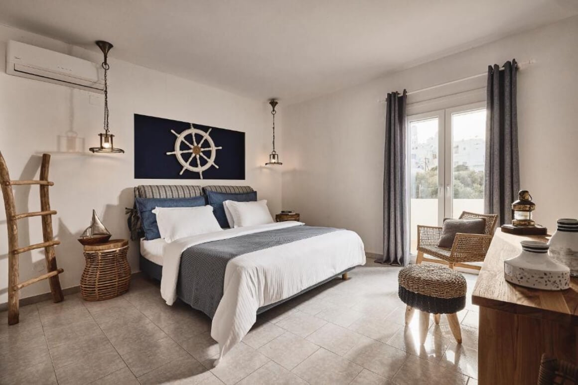 Sailor style bedroom with a queen bed with blue and white sheets. There is a canvas with a picture of a sailing boat steering wheel behind the bed.