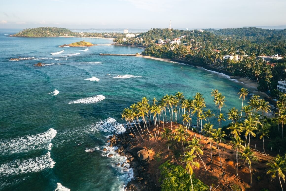 An aerial view of a Coconut tree hill in Mirissa beach with tall palm trees swaying in the breeze. 