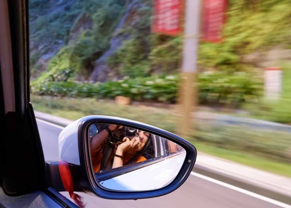 A girl takes a photo in the wing mirror of a car while driving along a road