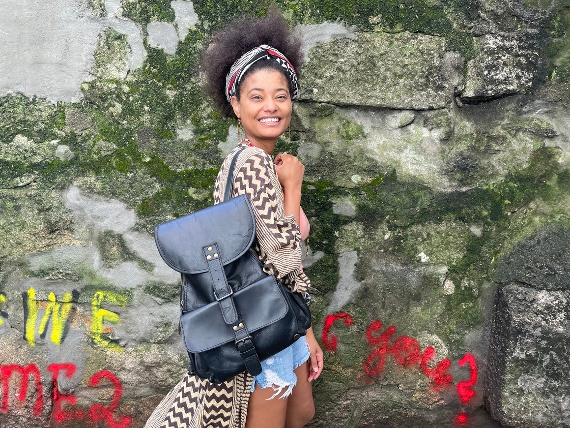 Girl standing by a wall with graffiti with a leather backpack on. 