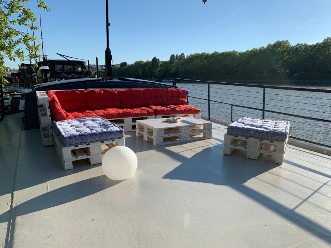 Terrace on top of the boat with pallet sofas and tables. View of the water and trees behind.