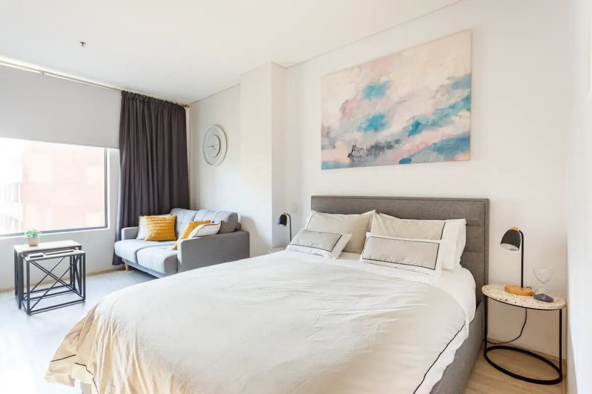 Clean, simple bedroom with a bed, sofa, coffee table, a large window and a piece of art behind the bed