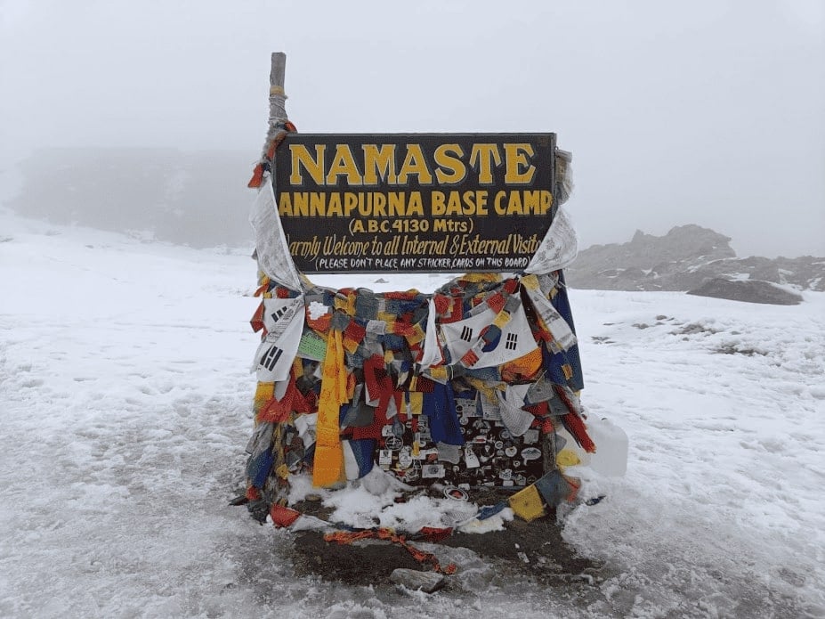 sign denoting annapurna basecamp with snow in the background and yellow red blue and orange prayer flags strung around it