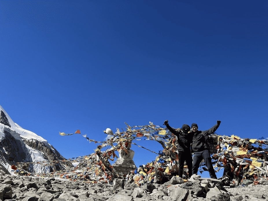 two guys standing in front of a ton of blowing prayer flags a top a mountain pass in nepal