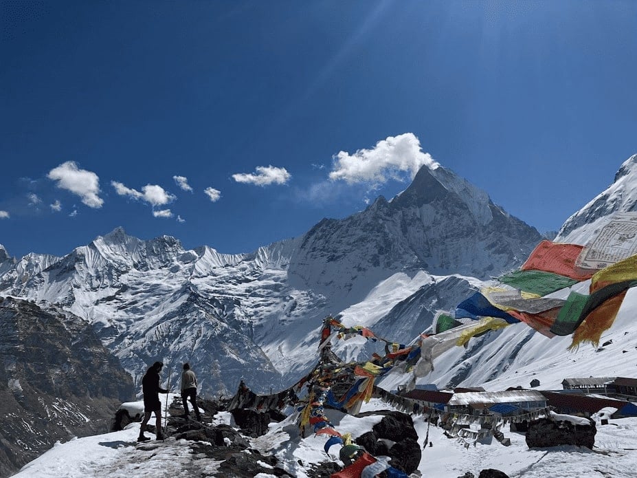 two people standing before a massive snow covered mountain while trekking the annapurna circuit in nepal with prayer flags flapping around them