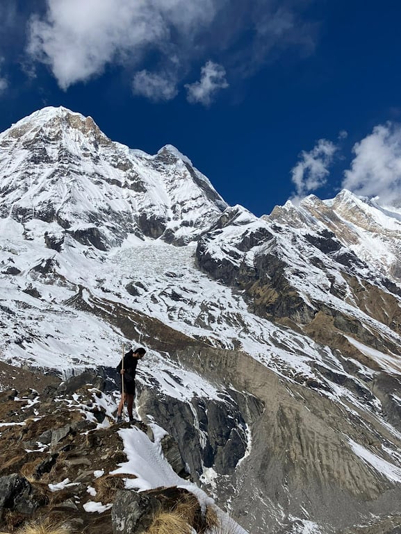 man holding a wooden stick standing on the edge of a snowcapped cliff while trekking in nepal