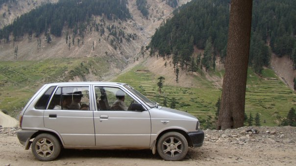 an old silver car driving in pakistan with three passengers in front of a large tree