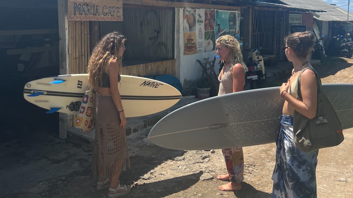 A group of friends talking outside a cafe holding surfboards.
