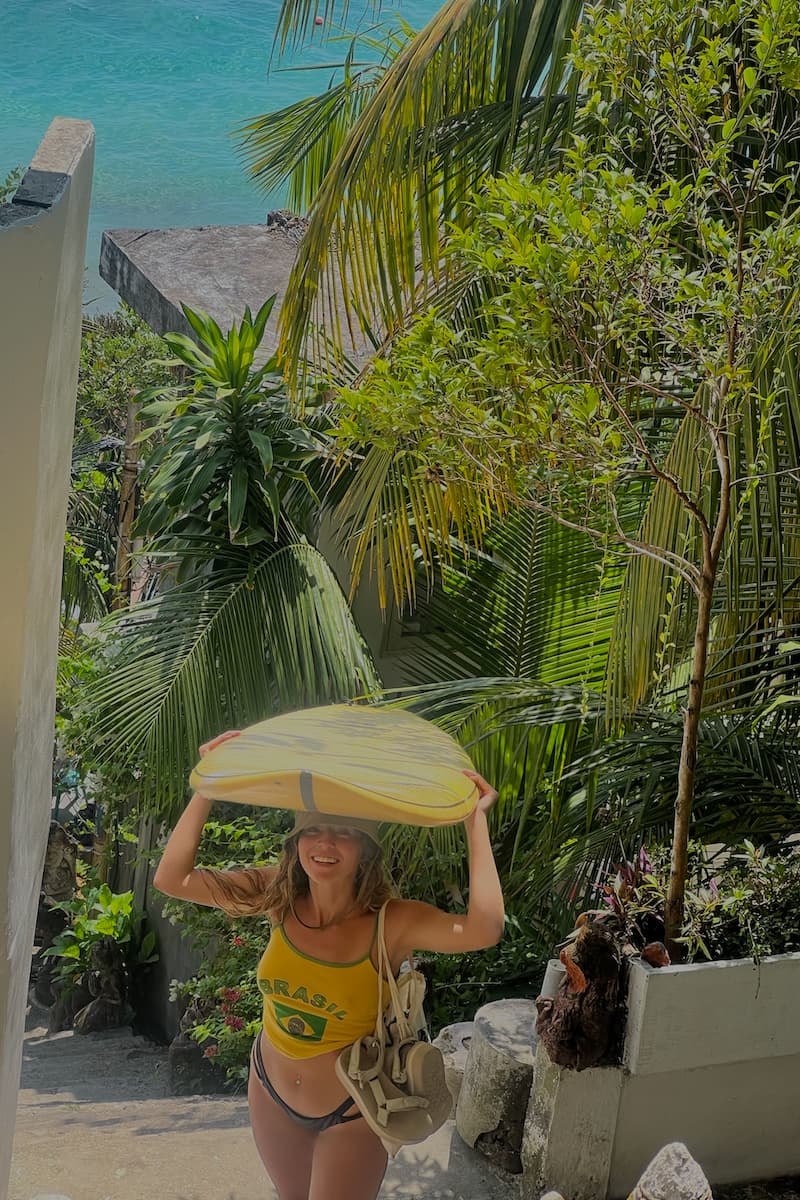 a girl holding a surfboard on top of her head as she walks up the stairs with the beach and palm trees in the background