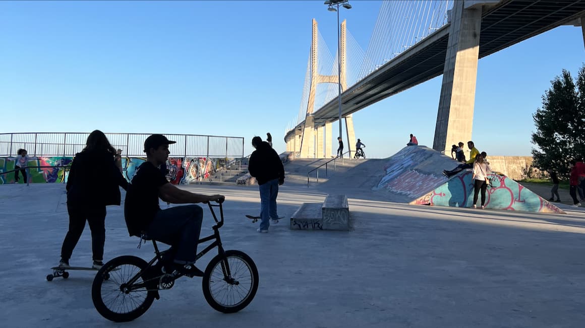 a few kids at a skatepark under a bridge one with a skateboard,and one riding a bike 