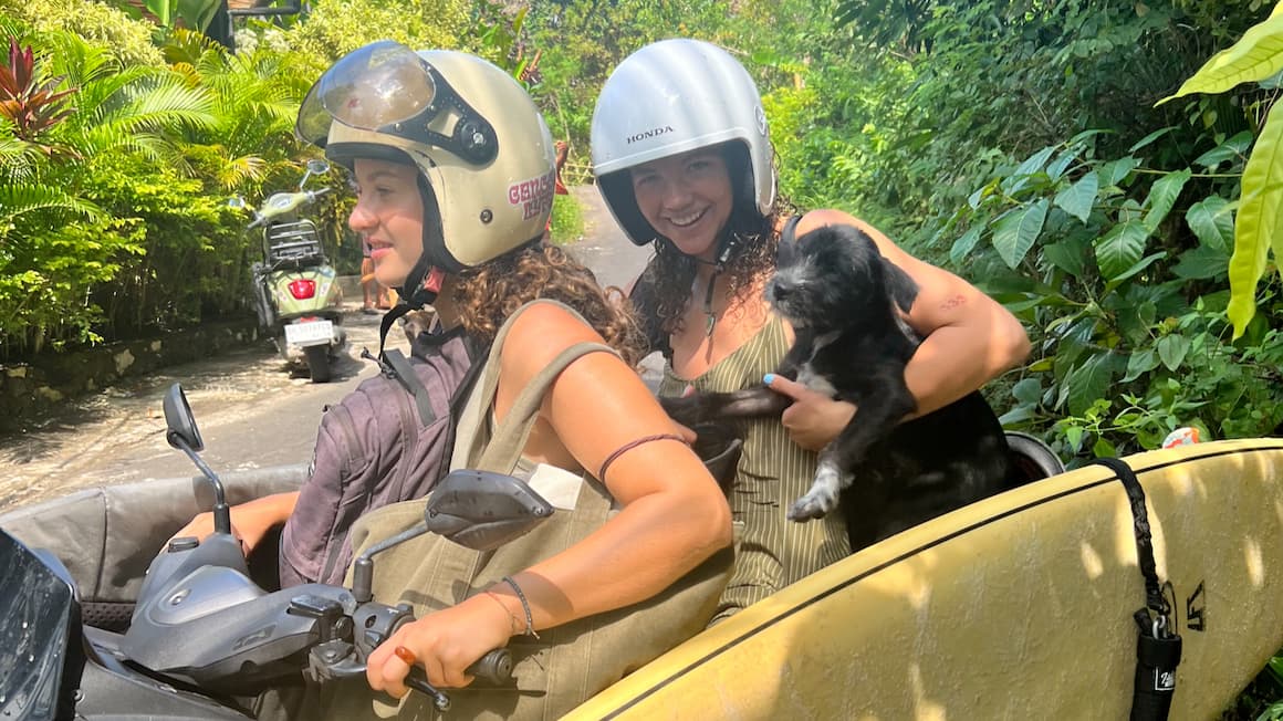 two girls on a scooter in Bali, Indonesia holding a small dog and with a surfboard hanging on the side of the scooter
