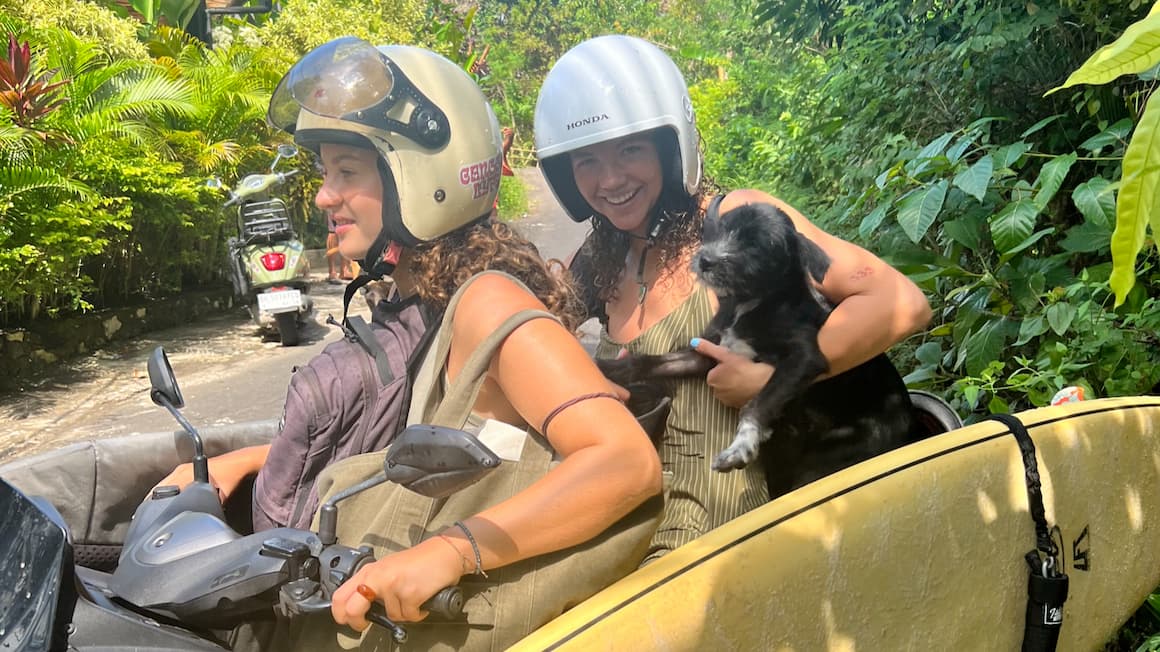 two girls on a scooter in Bali, Indonesia holding a small dog and with a surfboard hanging on the side of the scooter 