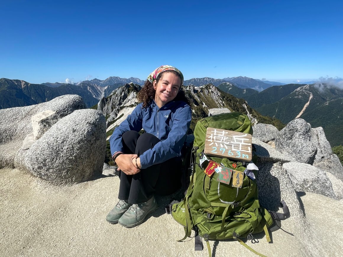 A girl sitting on top of a mountain cliff with her green Aether Backpack