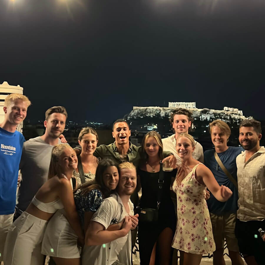 big group of friends on a rooftop in athens at night with view of the acropolis lit up in the background