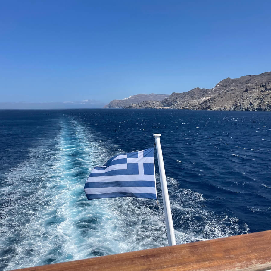Photo looking behind a boat over the ocean showing a greek flag waving behind the boat.