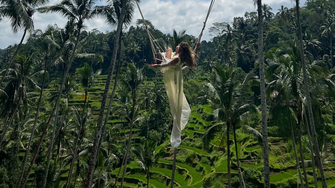 a girl on a swing with a white dress on and a flower in her hand with rice fields and palm trees in the background