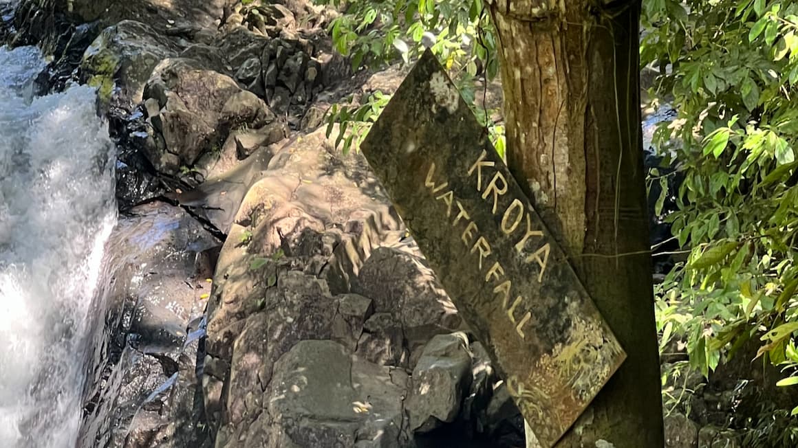 a sign that says Kroya waterfall, with the waterfall in the background