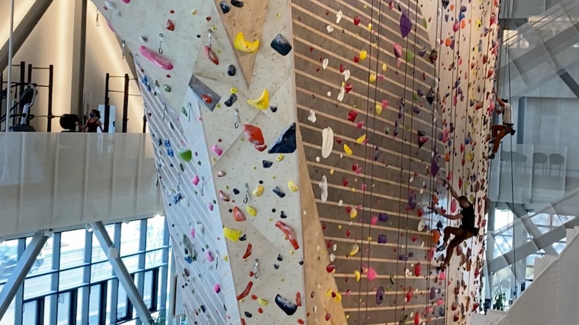 A guy rock climbing indoors, with a gym in the background