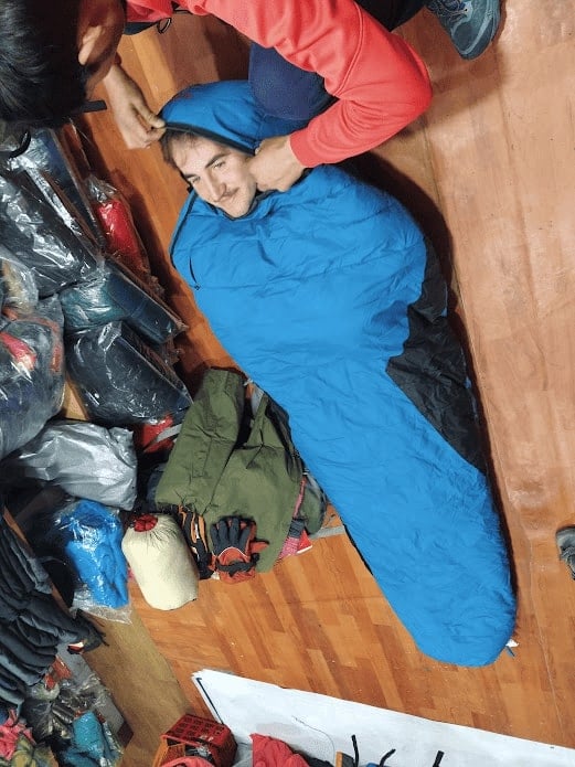 man laying in a bright blue sleeping bag on a wooden floor of a trekking gear shop in nepal