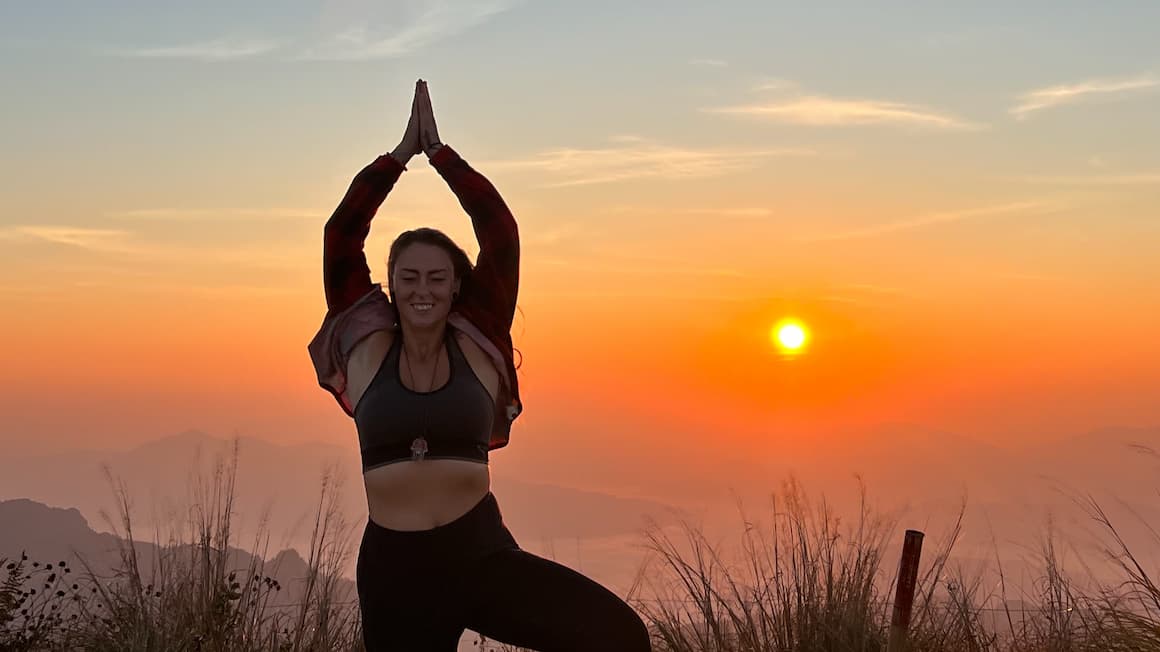a girl with her hands together in the sky holding a namaste yoga pose while the sun is rising