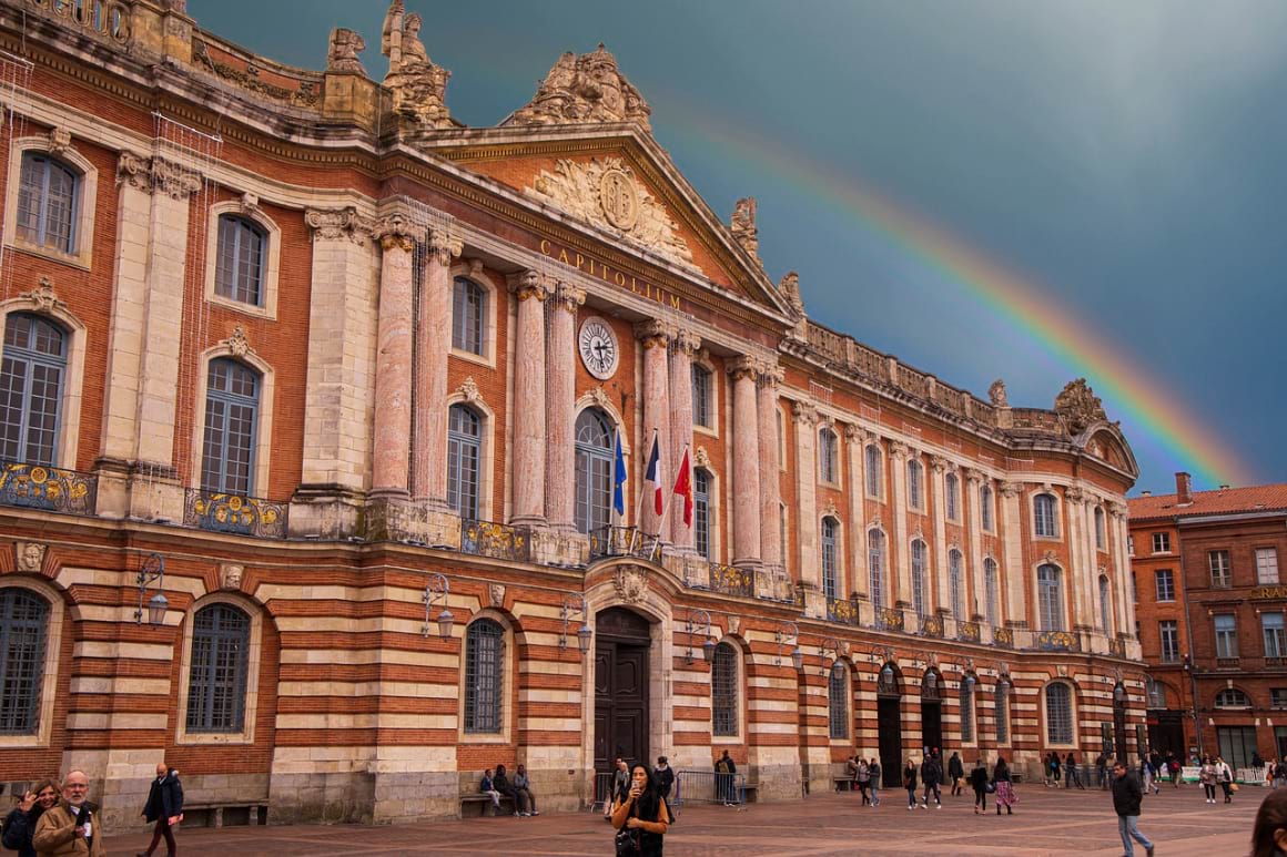 a bustling scene at Place du Capitole, with people strolling around its large courtyard and a rainbow in the sky