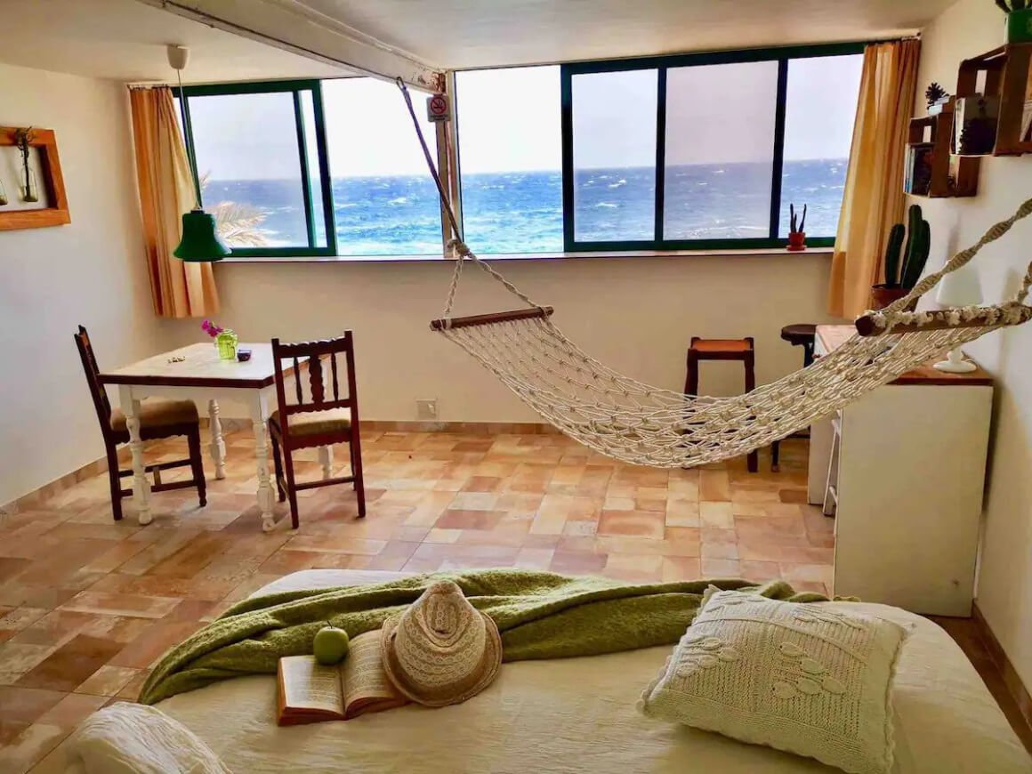 Room with bed, dining area, hammock and windows looking out directly to the ocean