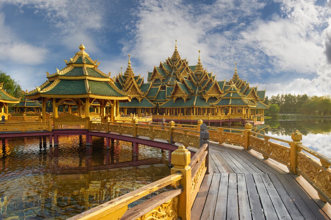 a wooden bridge leading to a traditional golden temple with green rooftop that's in the middle of a lake in Muang Boran Ancient City