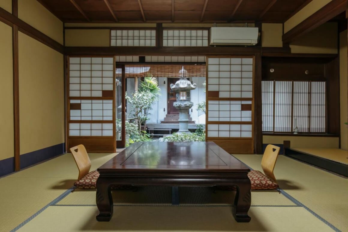 a room in Toshiharu Ryokan with a table and chairs right in the middle, sliding paper doors and a view of a garden.
