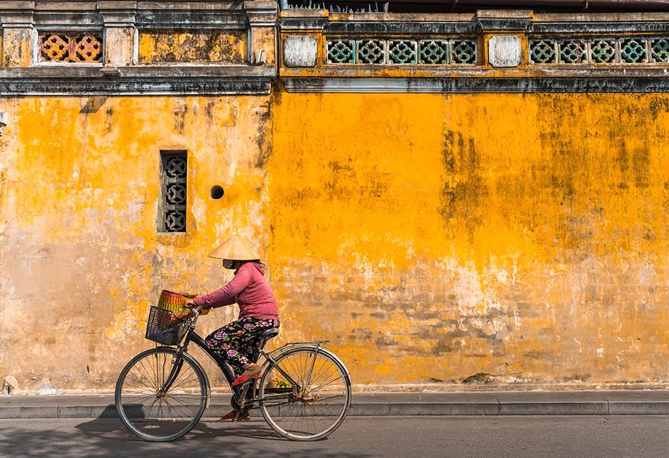 A woman on a bicycle with a vietnamese hat on riding past a yellow wall in Vietnam