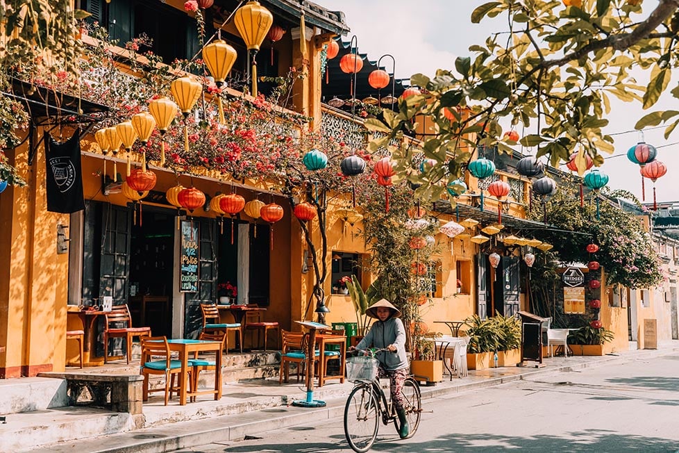 A person on a bicycle with a Vietnamese hat riding past a yellow house with colourful lanterns in Hoi An, Vietnam