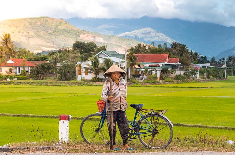 A woman standing next to her bike with a Vietnamese hat on next to a rice paddy with mountains in the distance in Vietnam