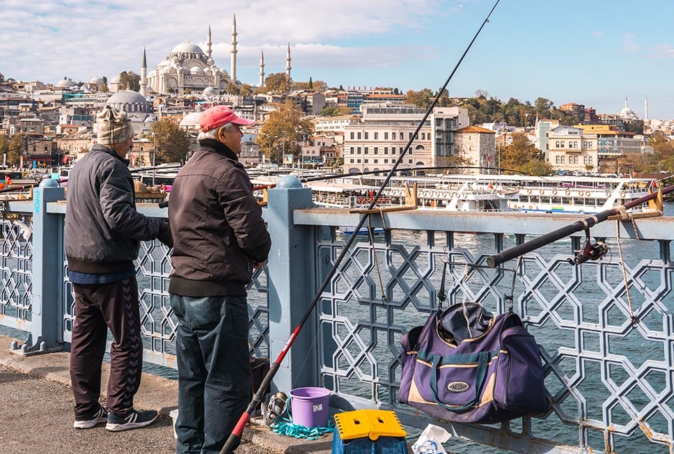 Two men fish from a bridge over the Bosphorus in Istanbul, Turkey with a large mosque in the background.
