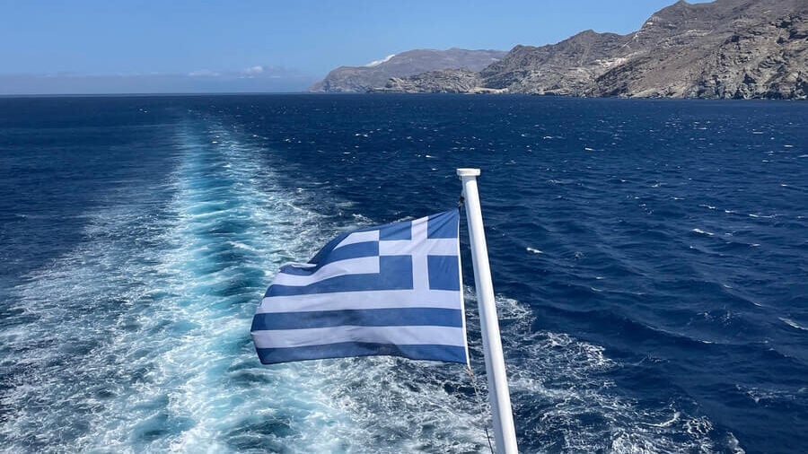 Photo looking behind a boat over the ocean showing a greek flag waving behind the boat.