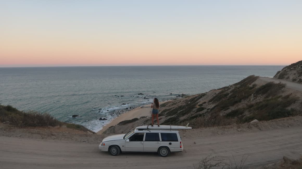 a girl standing on top of a white car as she looks at the sunset and the ocean
