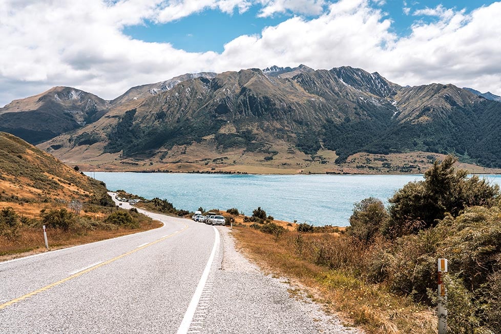 A road with a lake at the side of it and towering mountains in the background in Wanaka, New Zealand