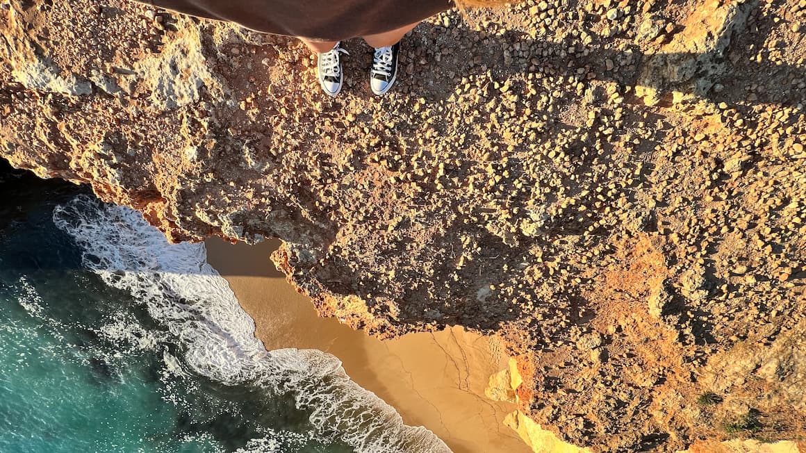 a girl standing on the edge of a cliff over looking a beach in portugal