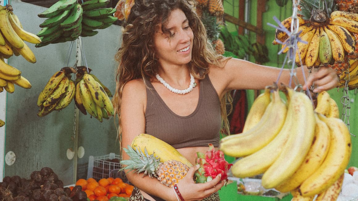 a girl at a fruit shop in Bali, Indonesia holding dragon fruit, a pineapple, and a papaya and picking bananas 