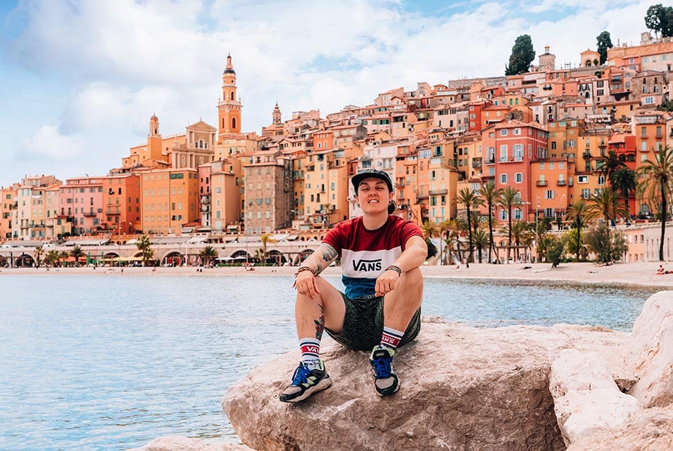 Nic sitting on a rock by the sea with the classic view of Menton, France behind them.