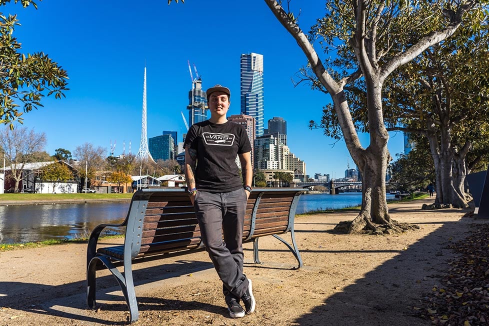 Nic leaning against a bench with the Melbourne skyline behind in Melbourne, Australia.