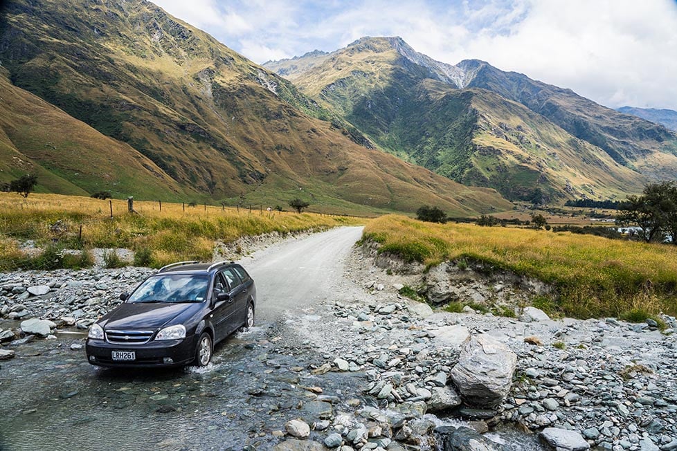 A car crossing a river on a dirt track with mountains in the distance near Queenstown, New Zealand
